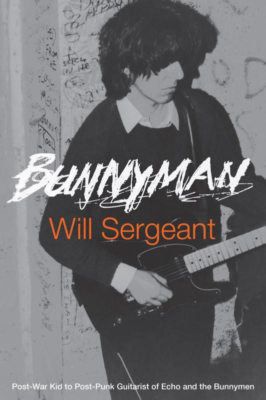 a black and white photo of a man with shaggy hair plays an electric guitar against a wall covered in writing 