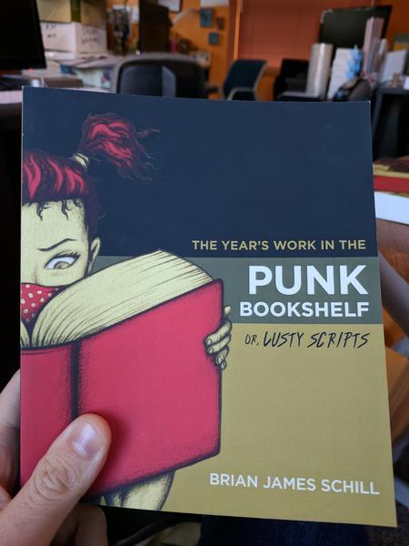 The Year's Work in the Punk Bookshelf or, Lusty Scripts by Brian James Schill (the cover has a young woman with red pigtails and a red bandana over her mouth, reading from a large red book)