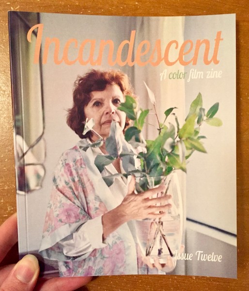 Set behind a pale backdrop, an elderly women in a floral bathrobe holds a vase of plants