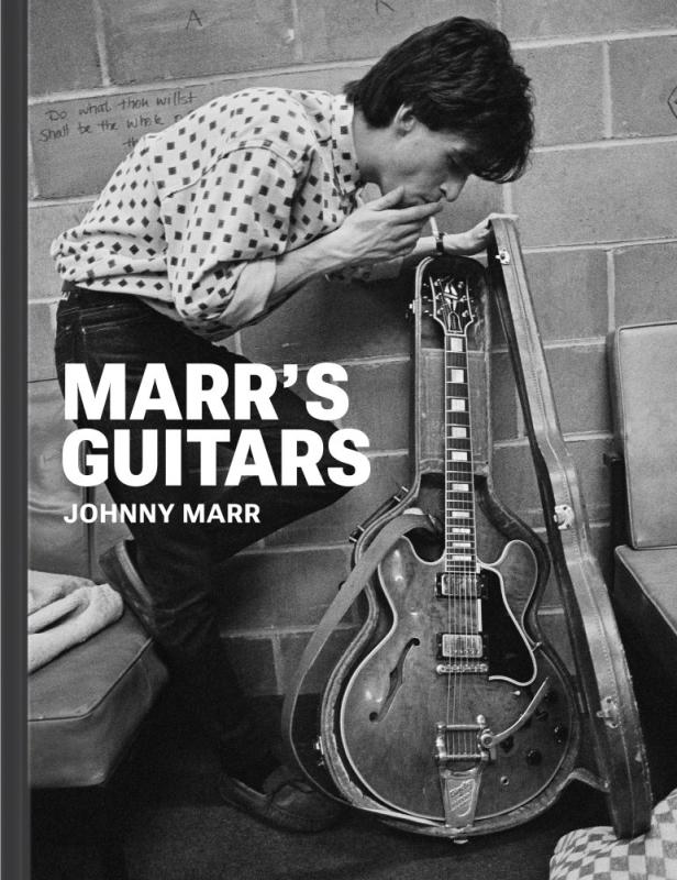 Black and white photo of Marr holding a Gibson hollow body electric guitar in a case, while smoking a cigarette, wearing a white and black polka dotted shirt.